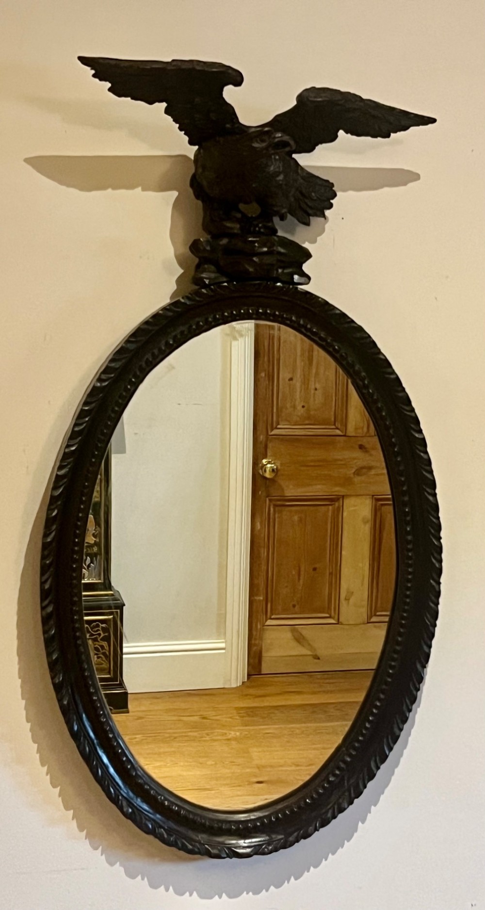 A dark stained oval wooden wall mirror featuring a carved eagle with spread wings perched atop a frame decorated with beaded and rope-twist detailing. View X