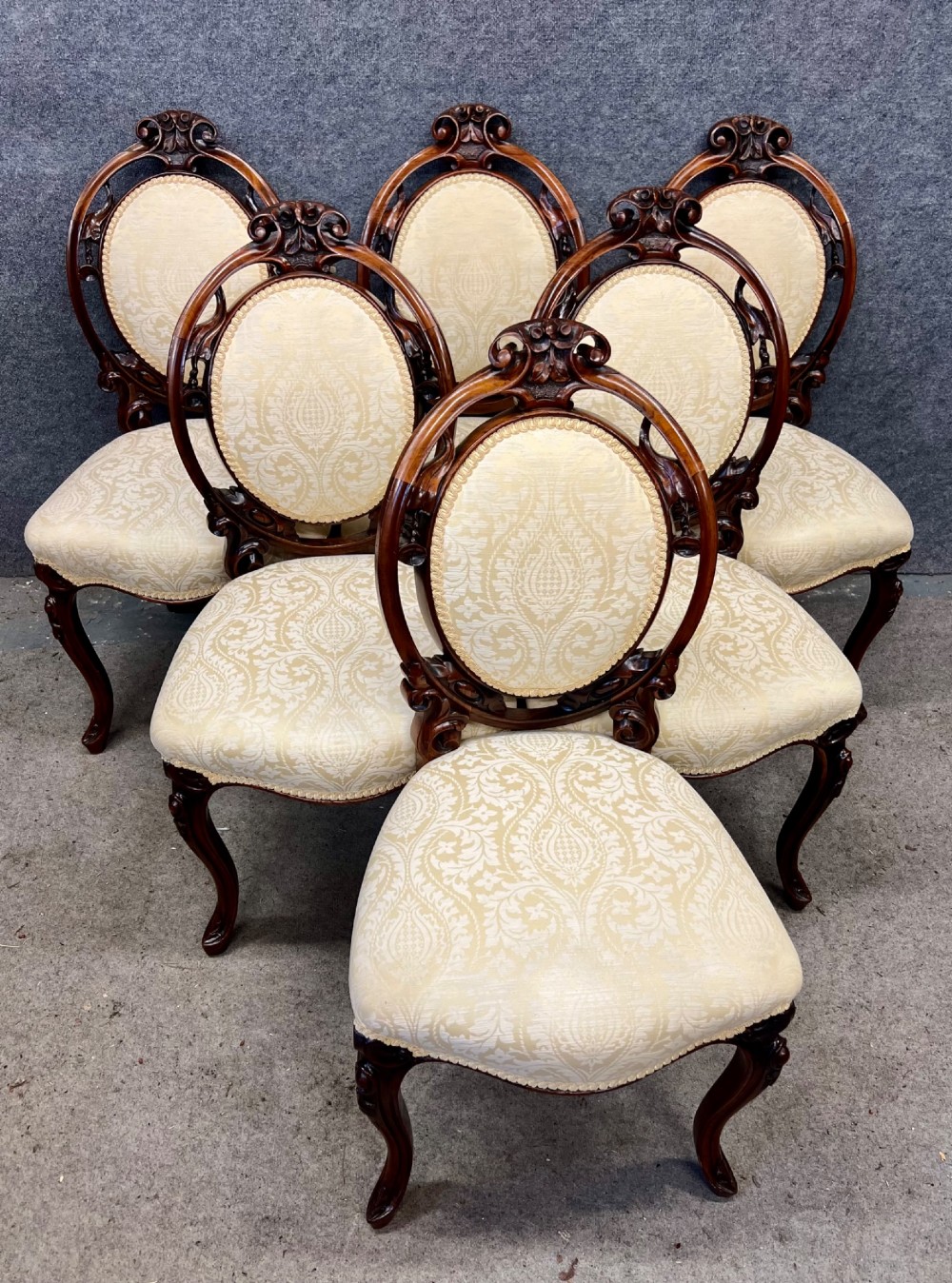 A set of six Victorian carved walnut dining chairs featuring oval balloon backs, cream patterned damask upholstery, and cabriole legs.