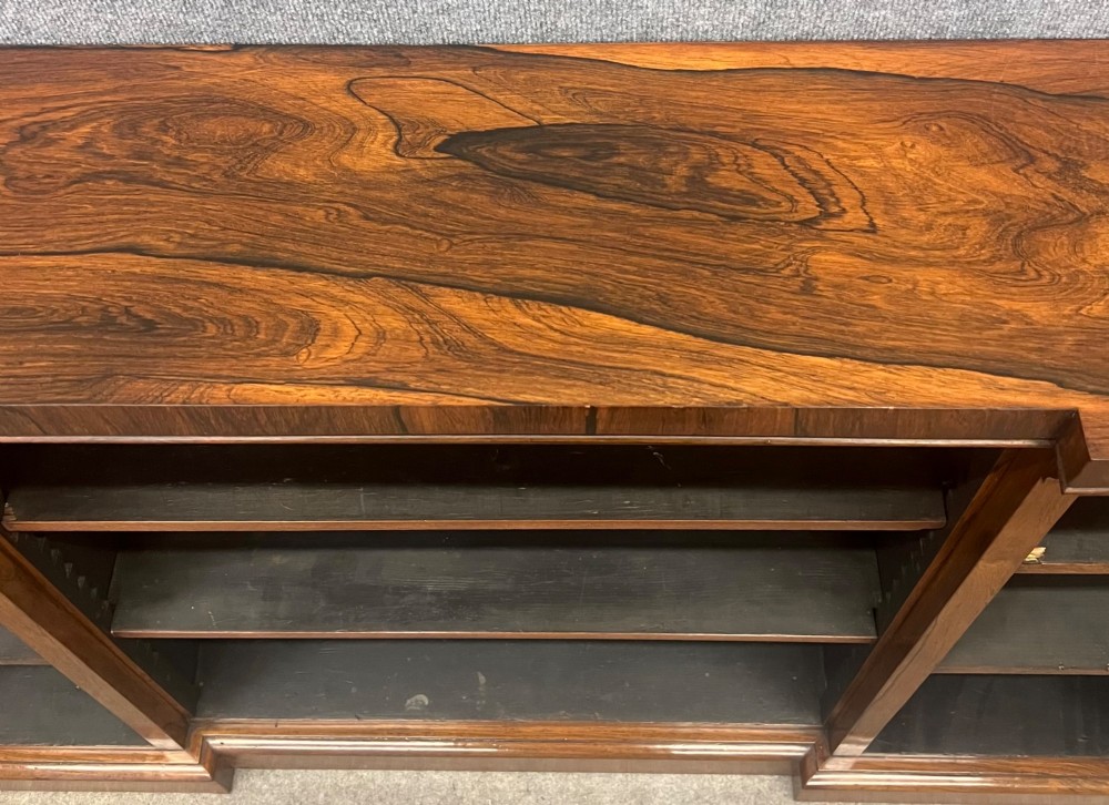 A low, wide Late Regency open rosewood bookcase featuring a breakfront top, three divided shelving sections, and a solid plinth base. View VI