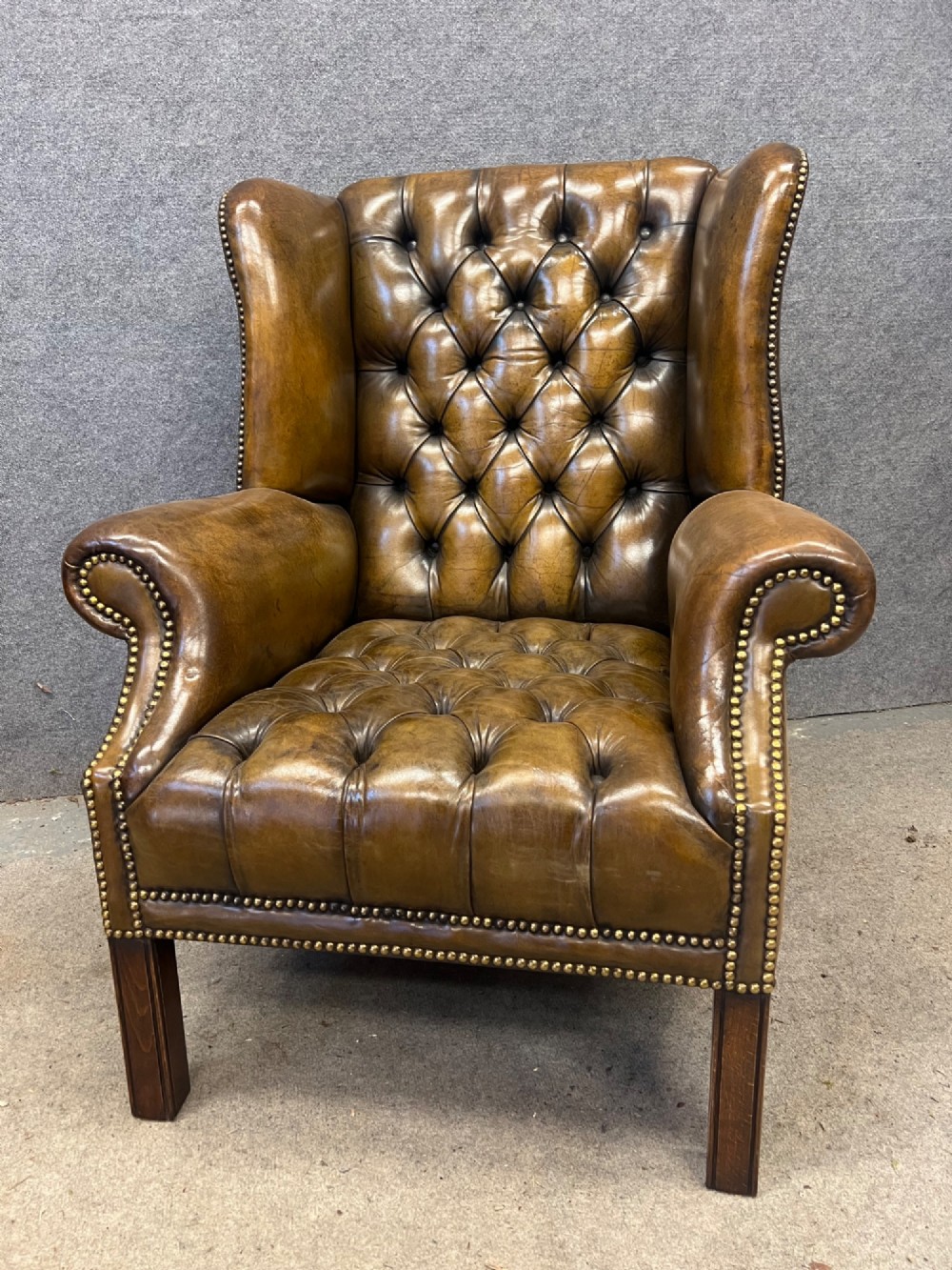 A Georgian style brown leather wingback armchair featuring deep buttoned tufting, rolled arms, brass stud detailing, and square wooden legs. View VII