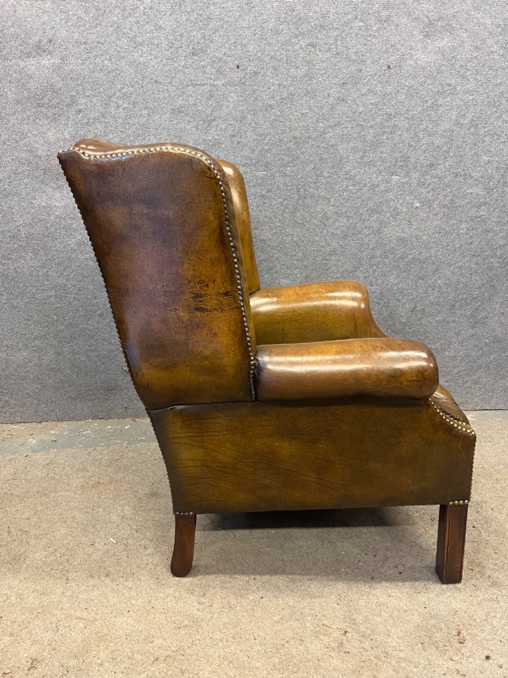 A Georgian style brown leather wingback armchair featuring deep buttoned tufting, rolled arms, brass stud detailing, and square wooden legs. View IX