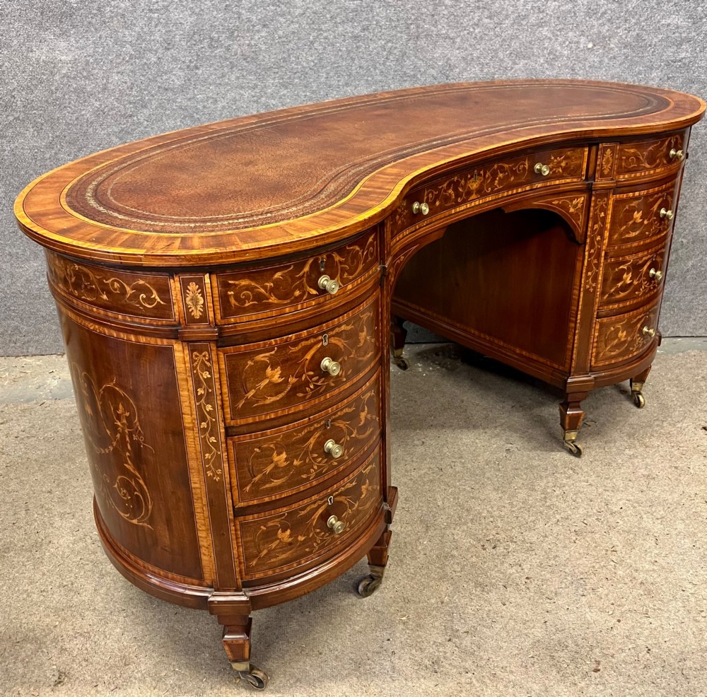 An Edwardian mahogany kidney-shaped desk featuring detailed floral marquetry inlay, a tooled leather writing surface, multiple drawers, and brass castors.
