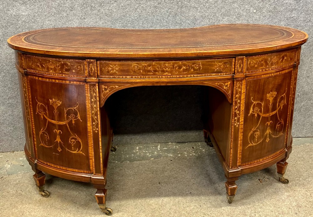 An Edwardian mahogany kidney-shaped desk featuring detailed floral marquetry inlay, a tooled leather writing surface, multiple drawers, and brass castors. View II