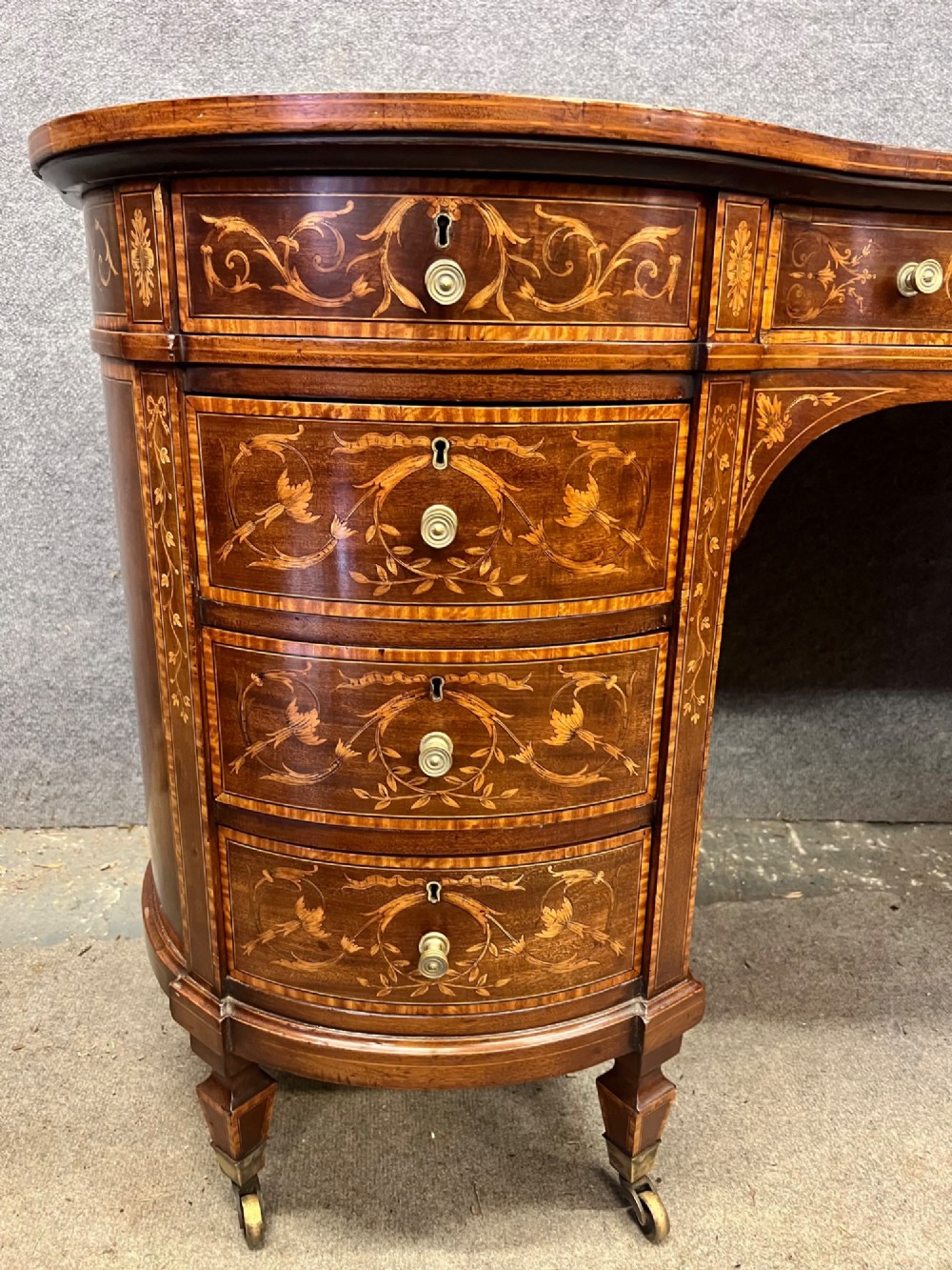 An Edwardian mahogany kidney-shaped desk featuring detailed floral marquetry inlay, a tooled leather writing surface, multiple drawers, and brass castors. View III