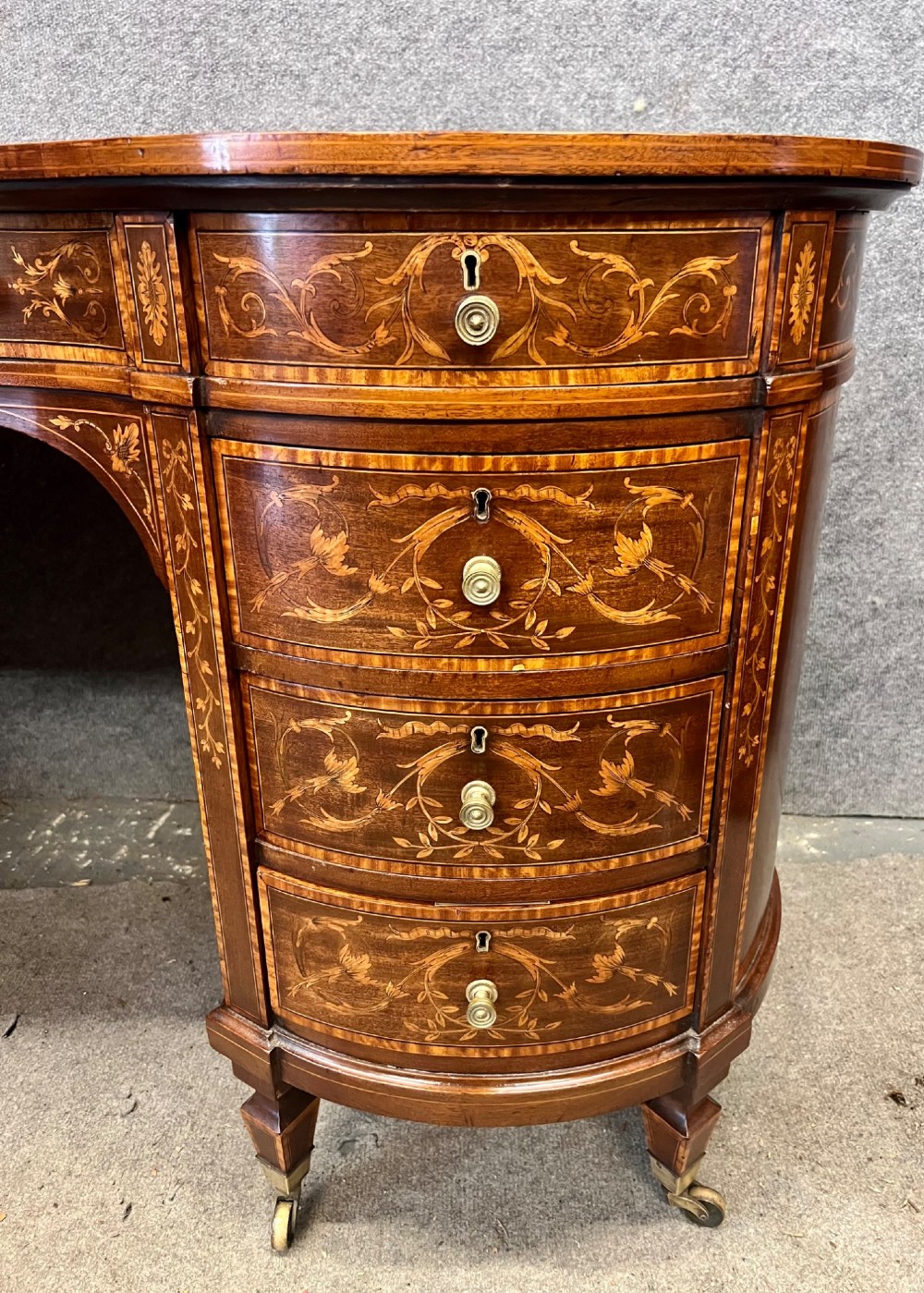 An Edwardian mahogany kidney-shaped desk featuring detailed floral marquetry inlay, a tooled leather writing surface, multiple drawers, and brass castors. View IV