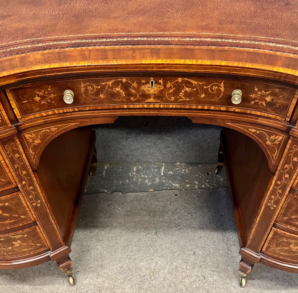 An Edwardian mahogany kidney-shaped desk featuring detailed floral marquetry inlay, a tooled leather writing surface, multiple drawers, and brass castors. View V