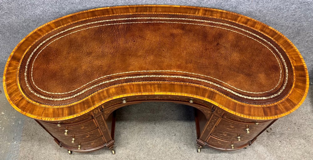 An Edwardian mahogany kidney-shaped desk featuring detailed floral marquetry inlay, a tooled leather writing surface, multiple drawers, and brass castors. View X