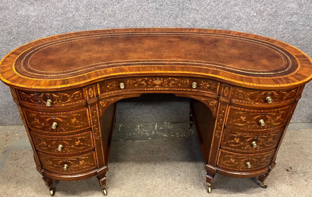 An Edwardian mahogany kidney-shaped desk featuring detailed floral marquetry inlay, a tooled leather writing surface, multiple drawers, and brass castors. View XI