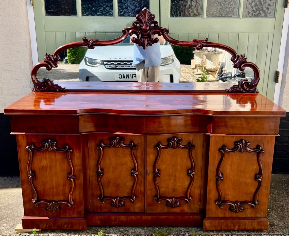 A nineteenth-century mahogany sideboard featuring a large carved mirror frame upstand, central double cupboard doors, flanking side doors with scroll mouldings, and flame timber veneers.