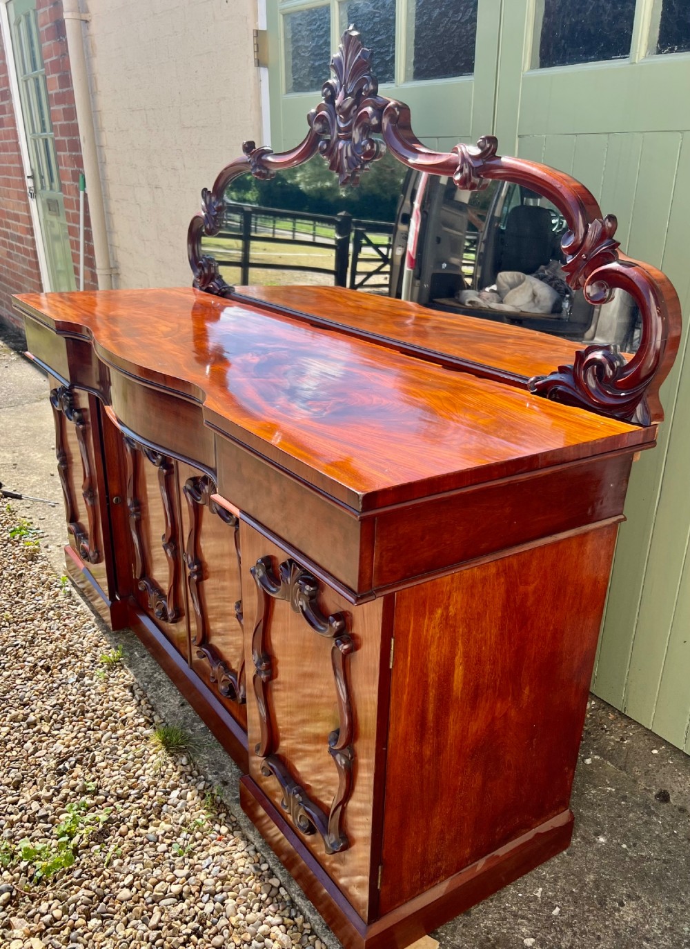 A nineteenth-century mahogany sideboard featuring a large carved mirror frame upstand, central double cupboard doors, flanking side doors with scroll mouldings, and flame timber veneers. View II