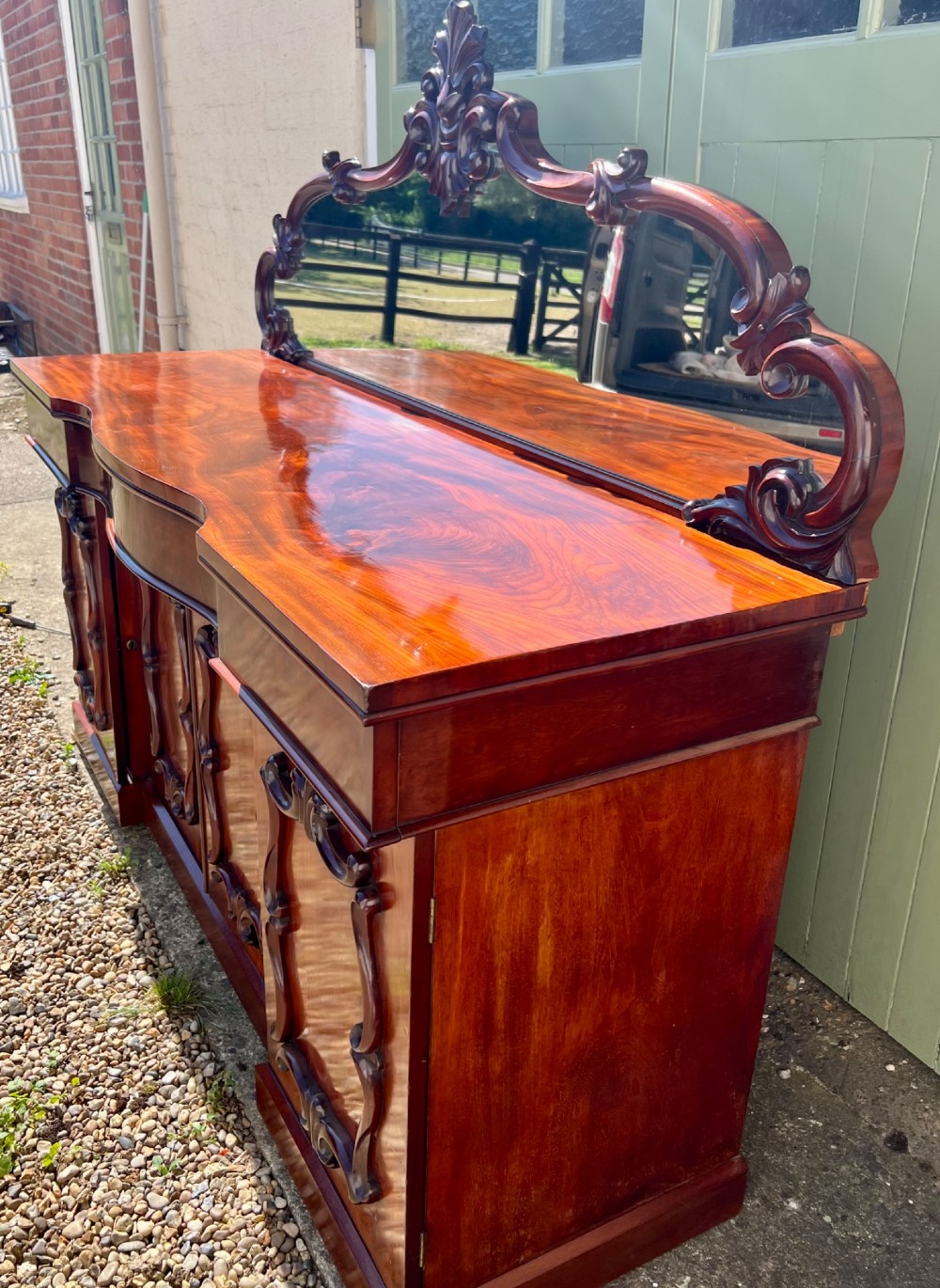 A nineteenth-century mahogany sideboard featuring a large carved mirror frame upstand, central double cupboard doors, flanking side doors with scroll mouldings, and flame timber veneers. View III