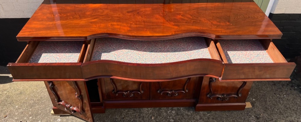 A nineteenth-century mahogany sideboard featuring a large carved mirror frame upstand, central double cupboard doors, flanking side doors with scroll mouldings, and flame timber veneers. View IX