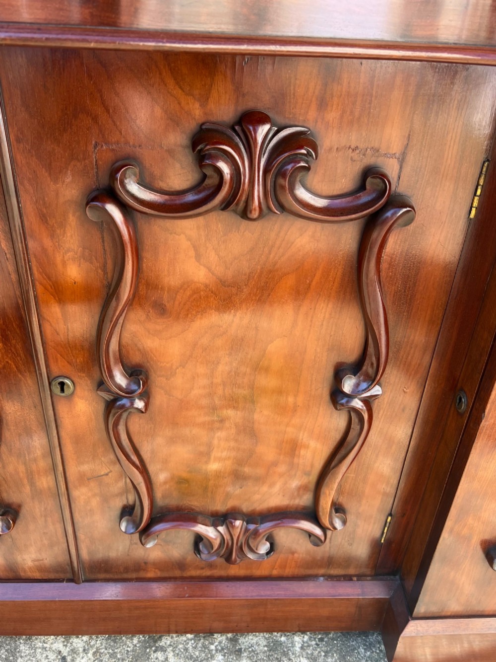 A nineteenth-century mahogany sideboard featuring a large carved mirror frame upstand, central double cupboard doors, flanking side doors with scroll mouldings, and flame timber veneers. View XI