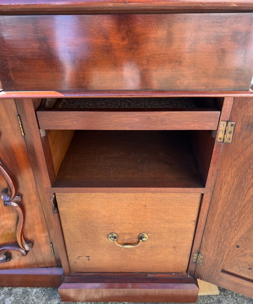 A nineteenth-century mahogany sideboard featuring a large carved mirror frame upstand, central double cupboard doors, flanking side doors with scroll mouldings, and flame timber veneers. View XV