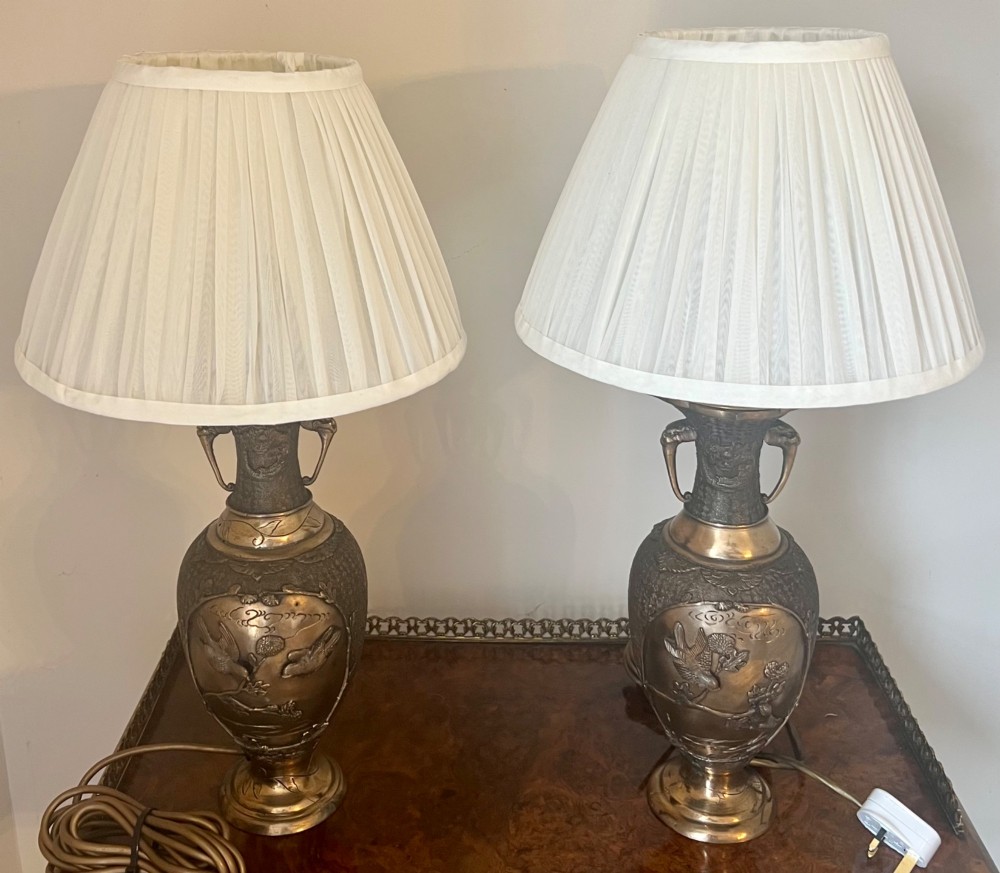 A pair of Japanese Meiji period bronze vase table lamps featuring relief bird and botanical motifs with pleated cream fabric lampshades.