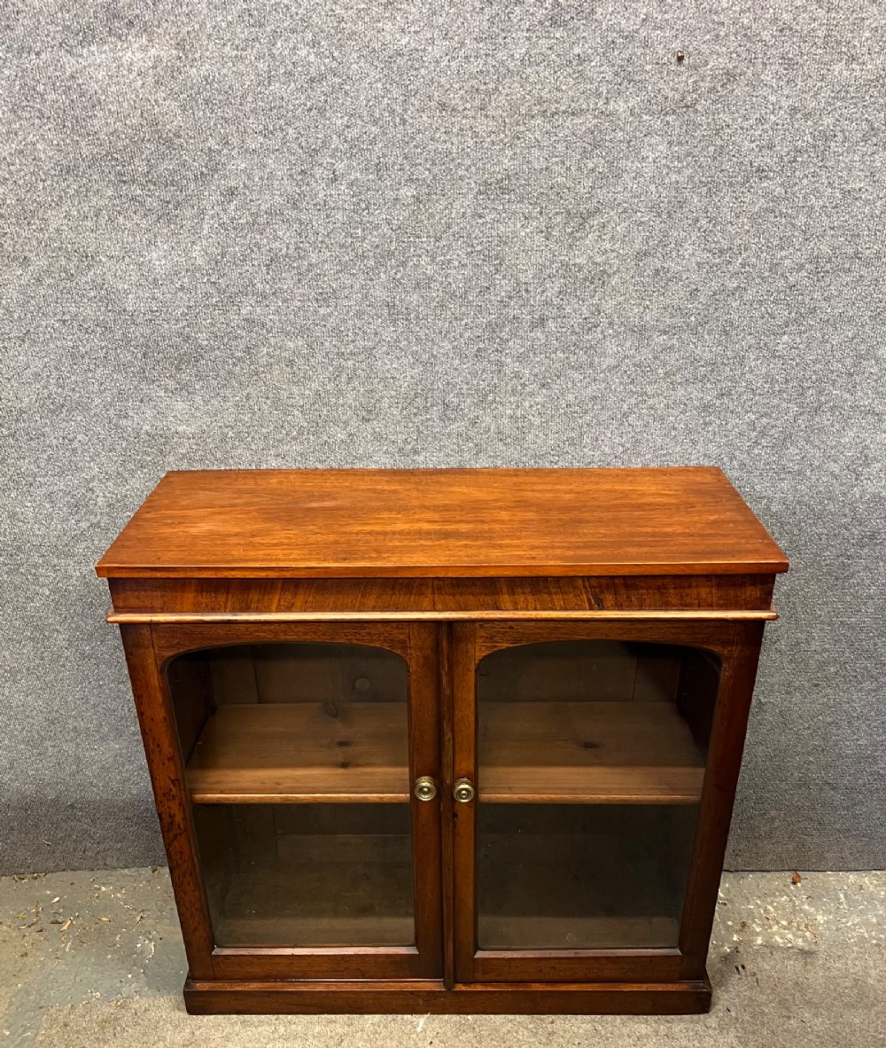 A small Victorian mahogany low bookcase featuring two glazed doors with arched frames, brass knobs, internal timber shelving, and a plinth base.