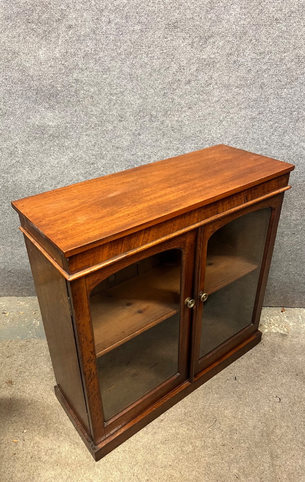 A small Victorian mahogany low bookcase featuring two glazed doors with arched frames, brass knobs, internal timber shelving, and a plinth base. View VI