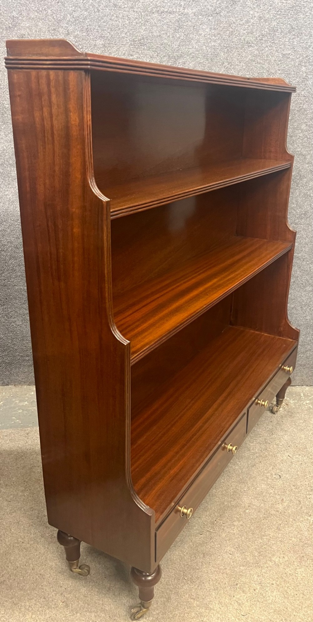 A warm mahogany open waterfall bookcase featuring three tiered shelves above twin drawers on turned legs with brass castors. View II
