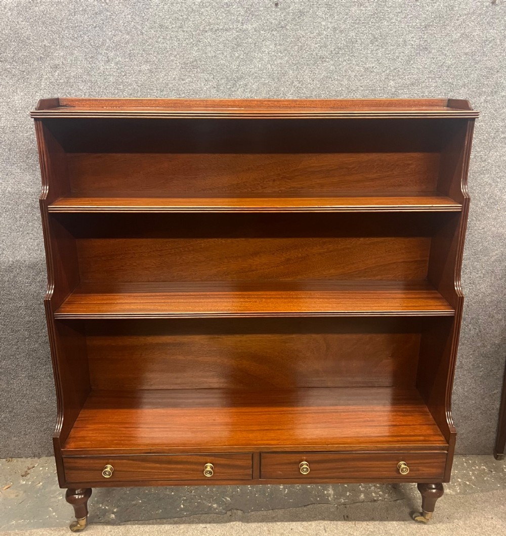 A warm mahogany open waterfall bookcase featuring three tiered shelves above twin drawers on turned legs with brass castors. View IX