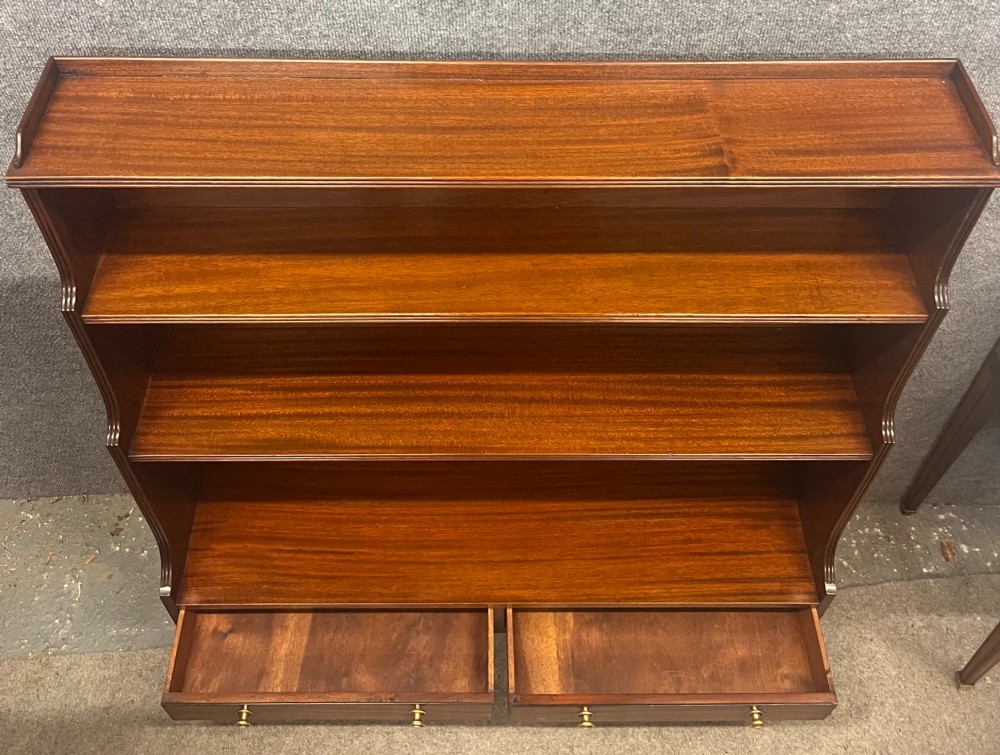 A warm mahogany open waterfall bookcase featuring three tiered shelves above twin drawers on turned legs with brass castors. View X