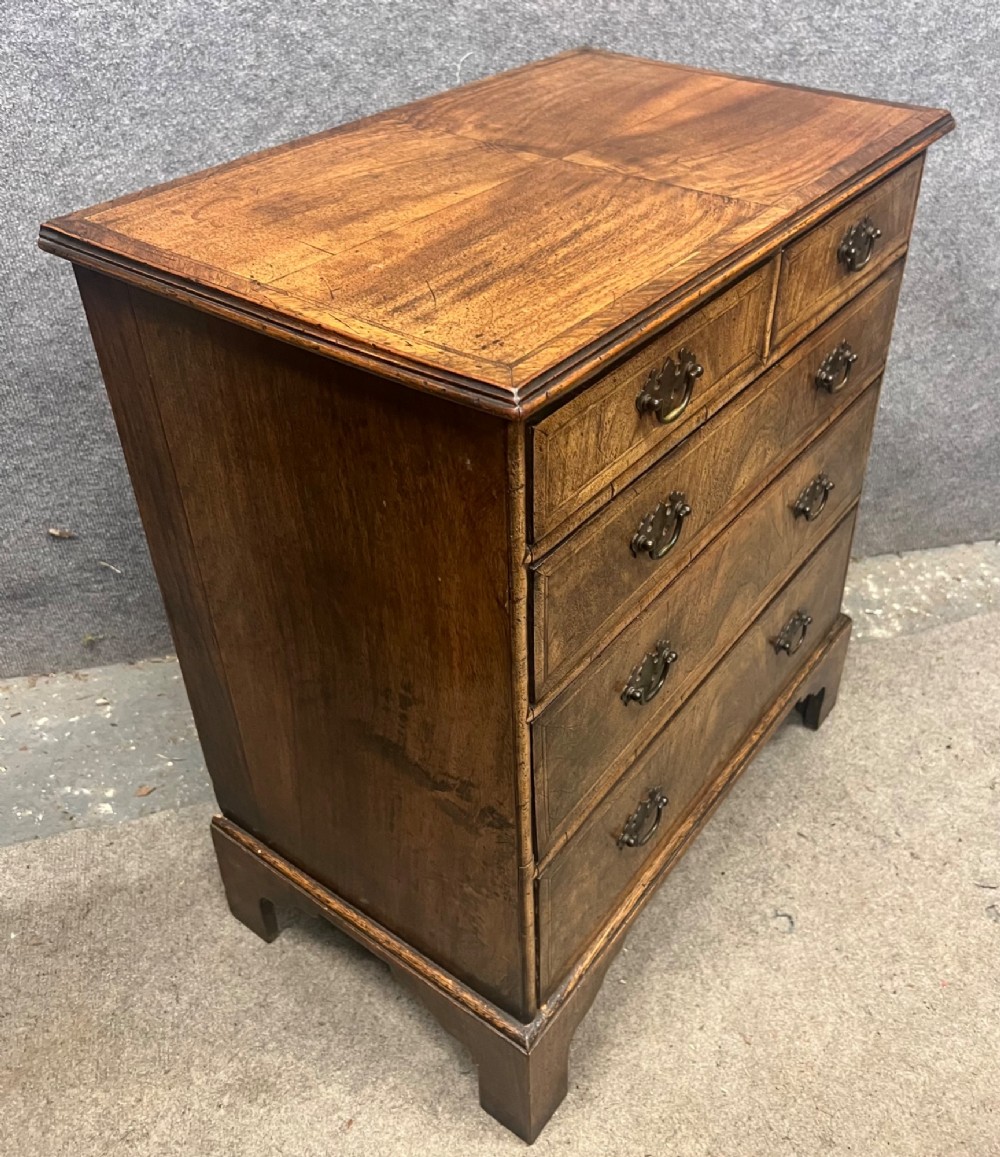 A small 18th-century style walnut chest of drawers featuring figured wood grain, brass drop handles, and bracket feet. View VII