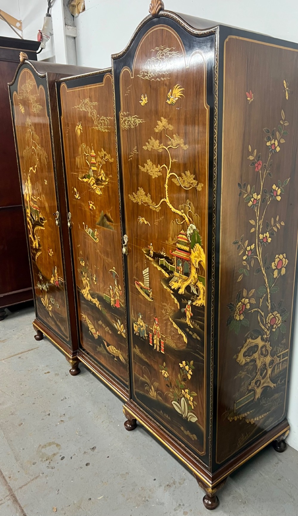 A triple-door Chinoiserie wardrobe featuring intricate gilded Asian landscape scenes, shell-carved crests, and dark polished wood frames. View IV
