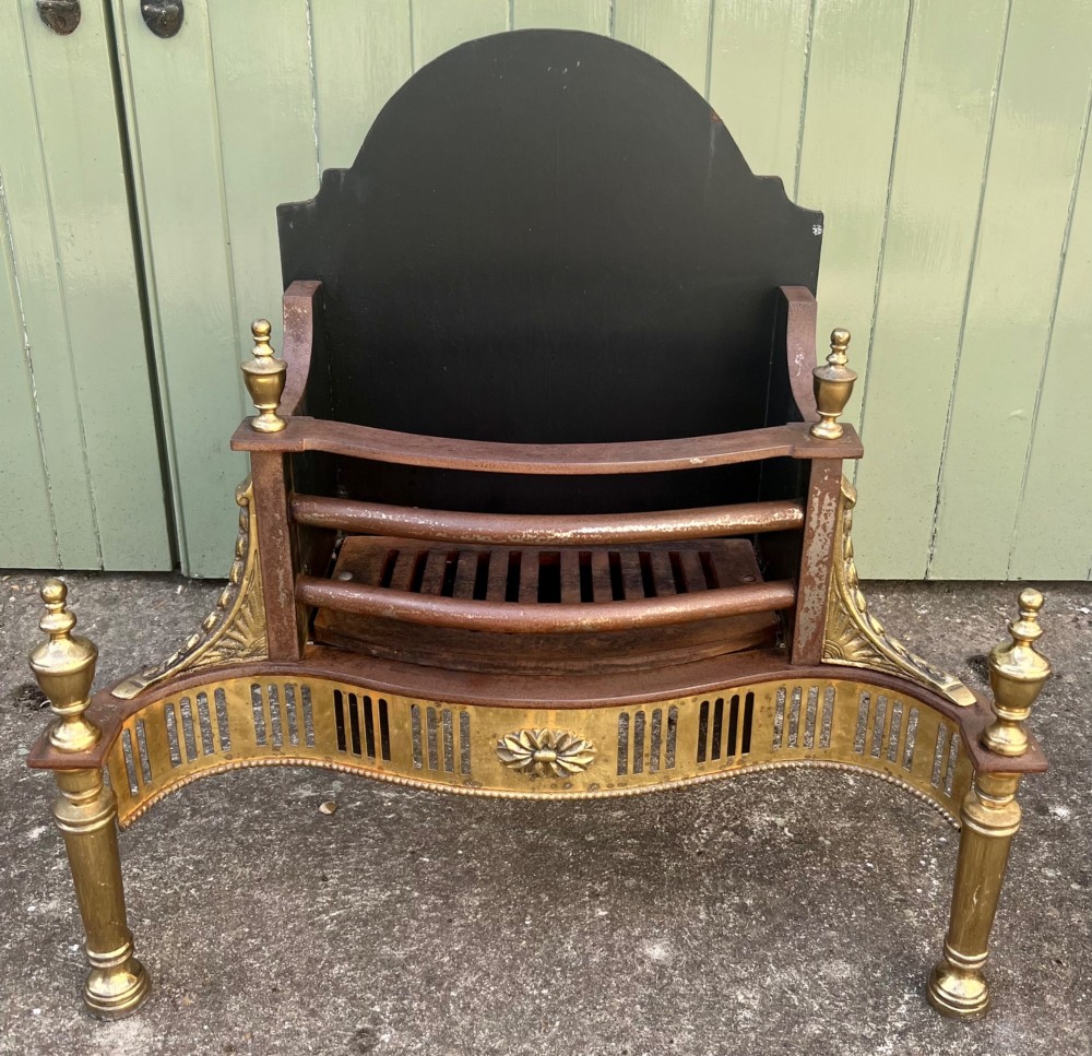 An Adam style brass and cast iron fire grate featuring a serpentine pierced front apron, urn-shaped finials, and a curved black backplate.