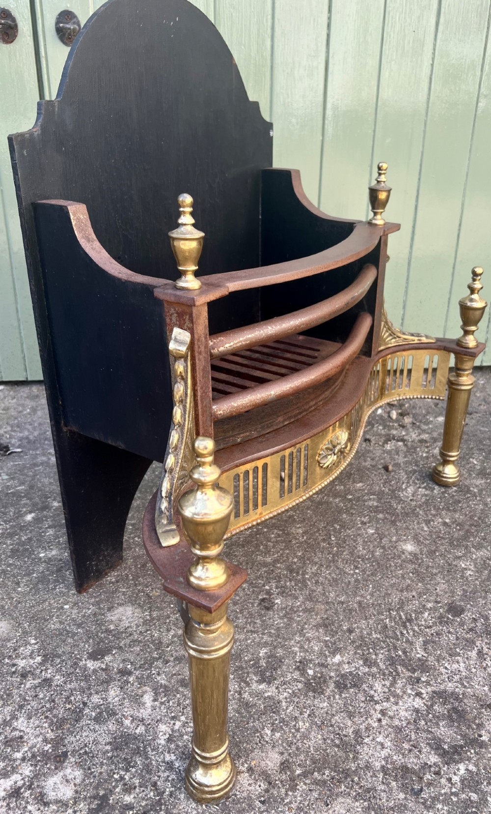 An Adam style brass and cast iron fire grate featuring a serpentine pierced front apron, urn-shaped finials, and a curved black backplate. View III