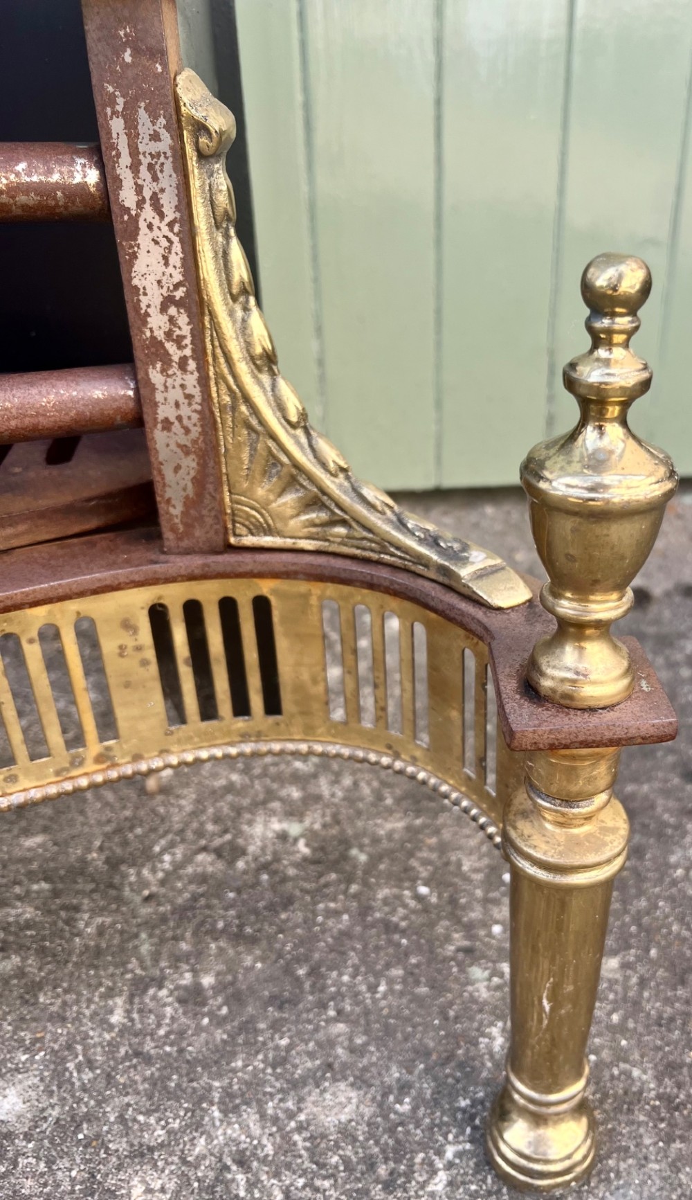 An Adam style brass and cast iron fire grate featuring a serpentine pierced front apron, urn-shaped finials, and a curved black backplate. View V