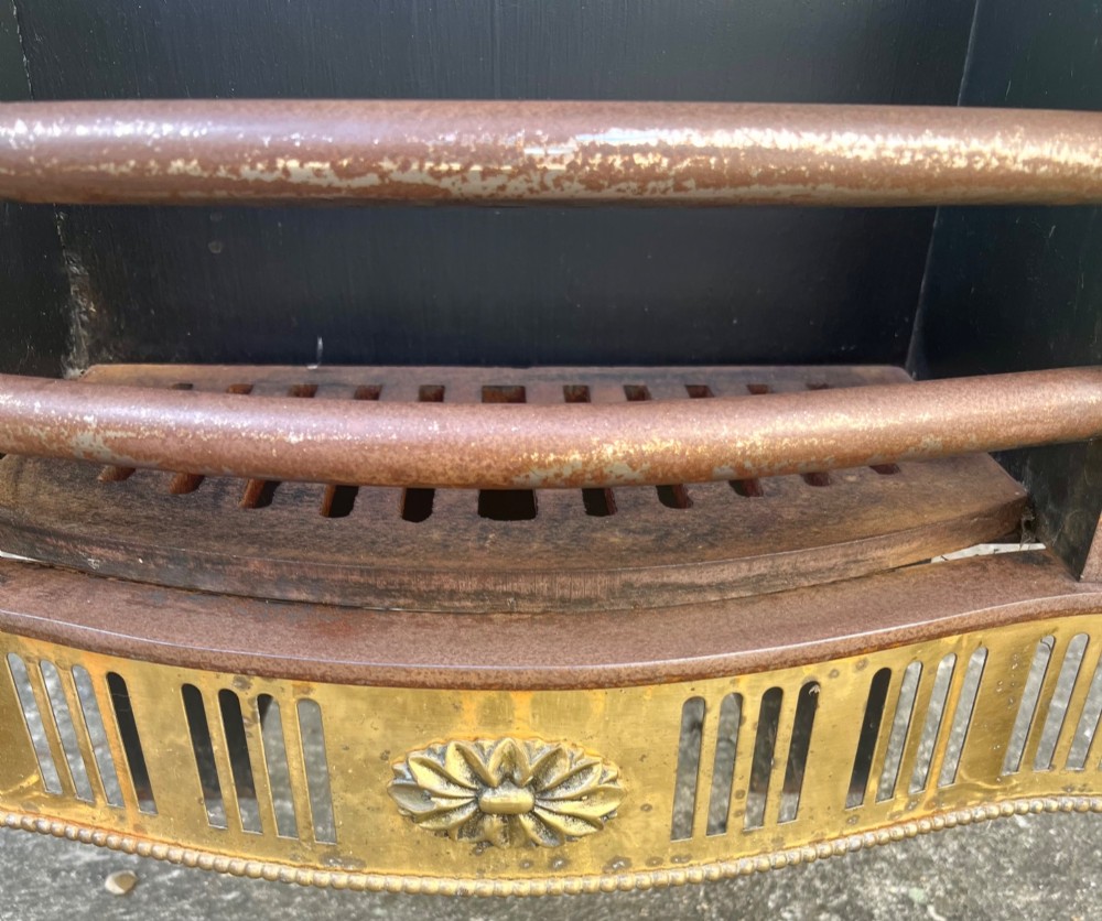 An Adam style brass and cast iron fire grate featuring a serpentine pierced front apron, urn-shaped finials, and a curved black backplate. View VI
