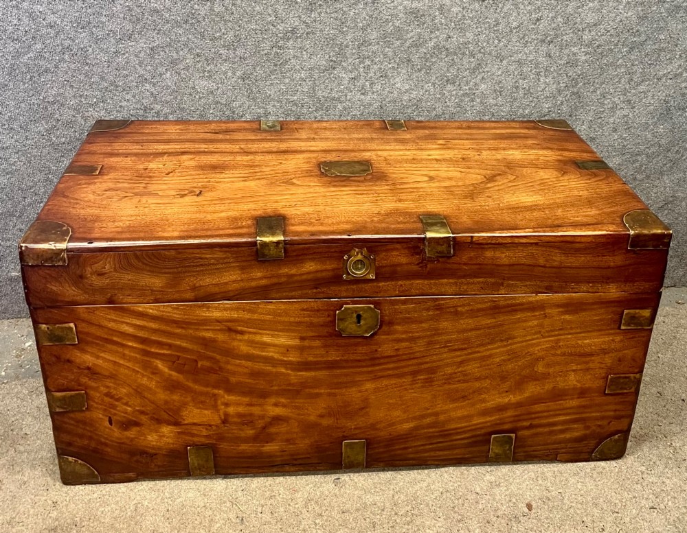 A rectangular warm brown wooden campaign trunk featuring flush brass corner brackets, metal straps, and a central brass lid plaque.