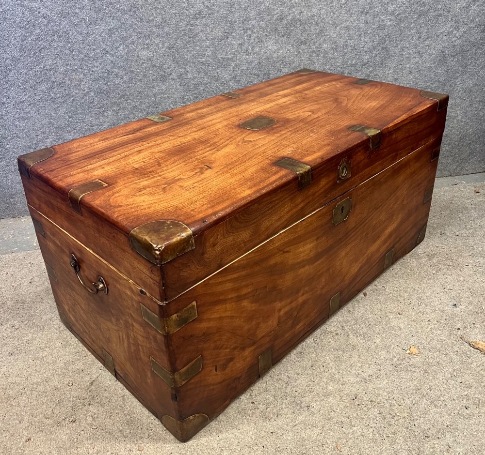 A rectangular warm brown wooden campaign trunk featuring flush brass corner brackets, metal straps, and a central brass lid plaque. View II