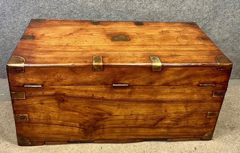 A rectangular warm brown wooden campaign trunk featuring flush brass corner brackets, metal straps, and a central brass lid plaque. View III