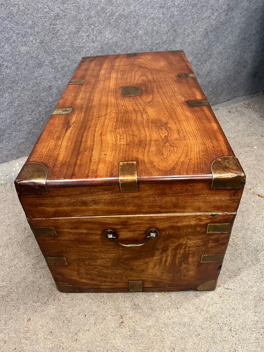 A rectangular warm brown wooden campaign trunk featuring flush brass corner brackets, metal straps, and a central brass lid plaque. View IV