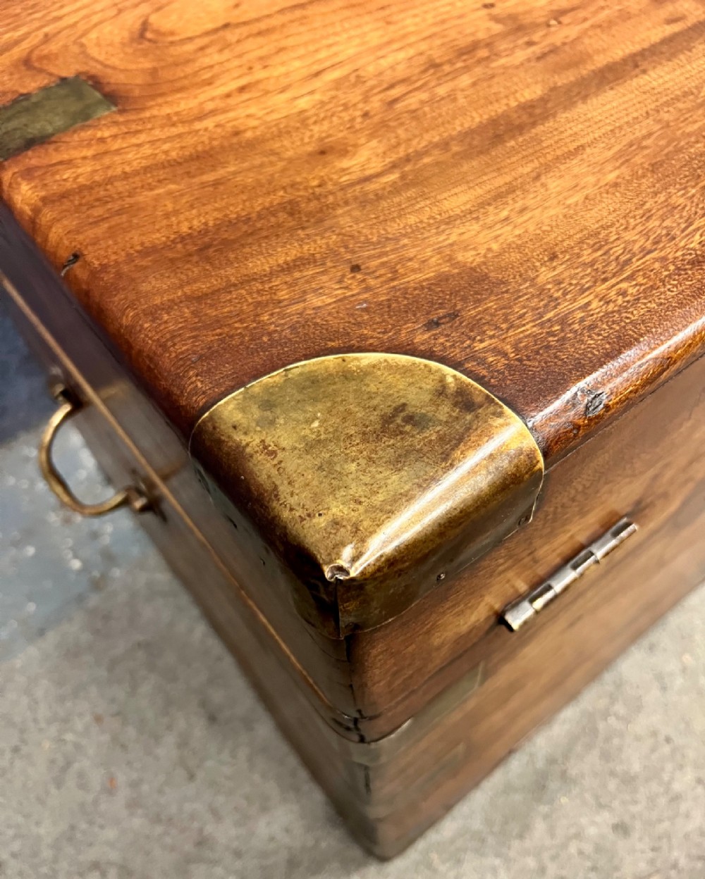 A rectangular warm brown wooden campaign trunk featuring flush brass corner brackets, metal straps, and a central brass lid plaque. View V