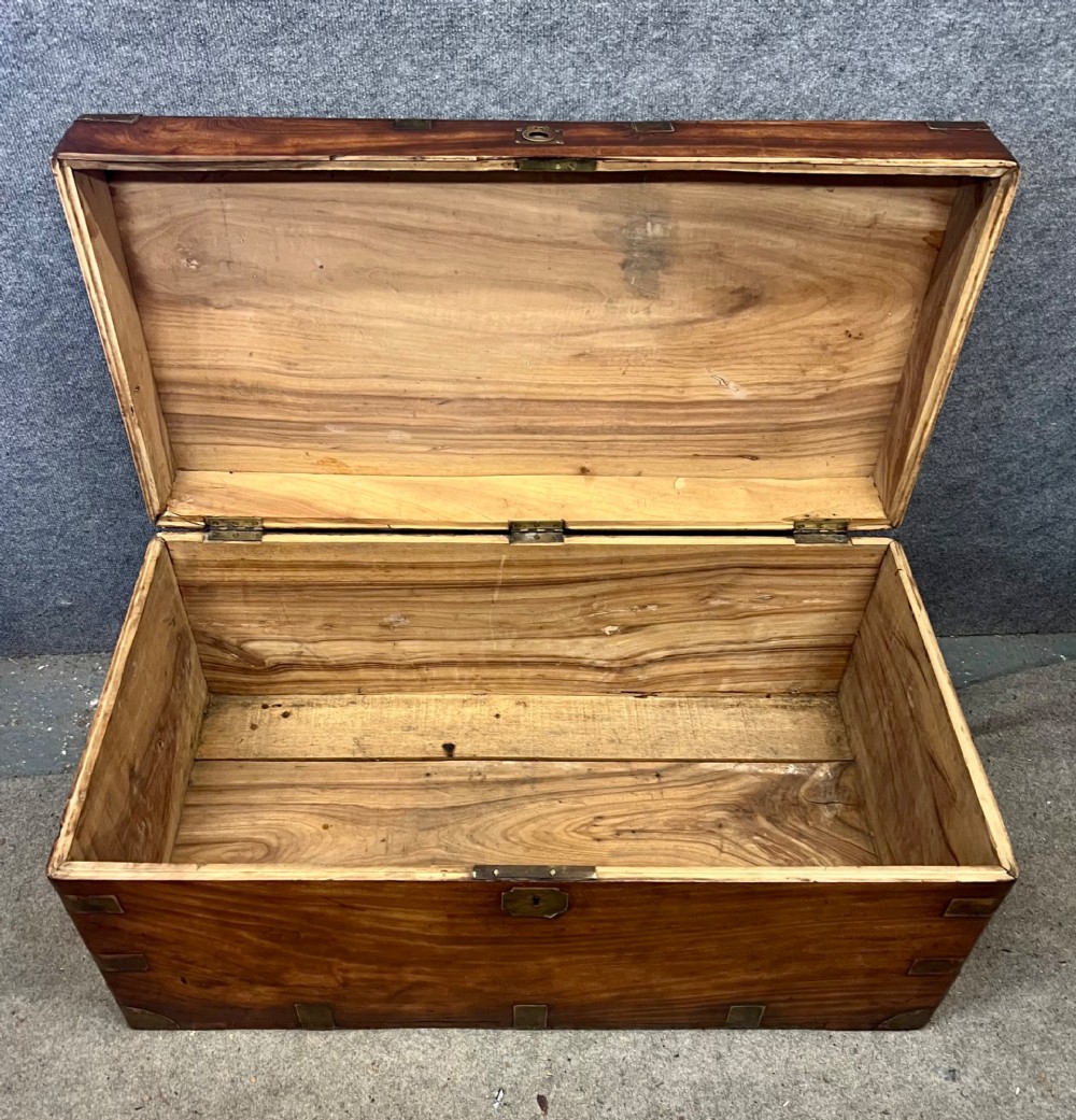 A rectangular warm brown wooden campaign trunk featuring flush brass corner brackets, metal straps, and a central brass lid plaque. View VII