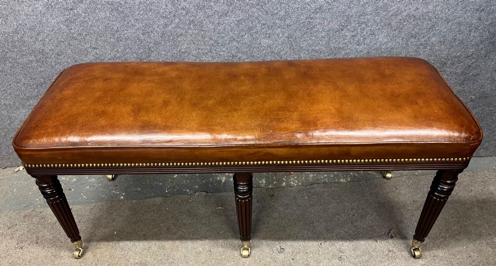 An early nineteenth-century upholstered brown leather bench featuring brass studded trim and six dark wood fluted turned legs on brass castors. View II