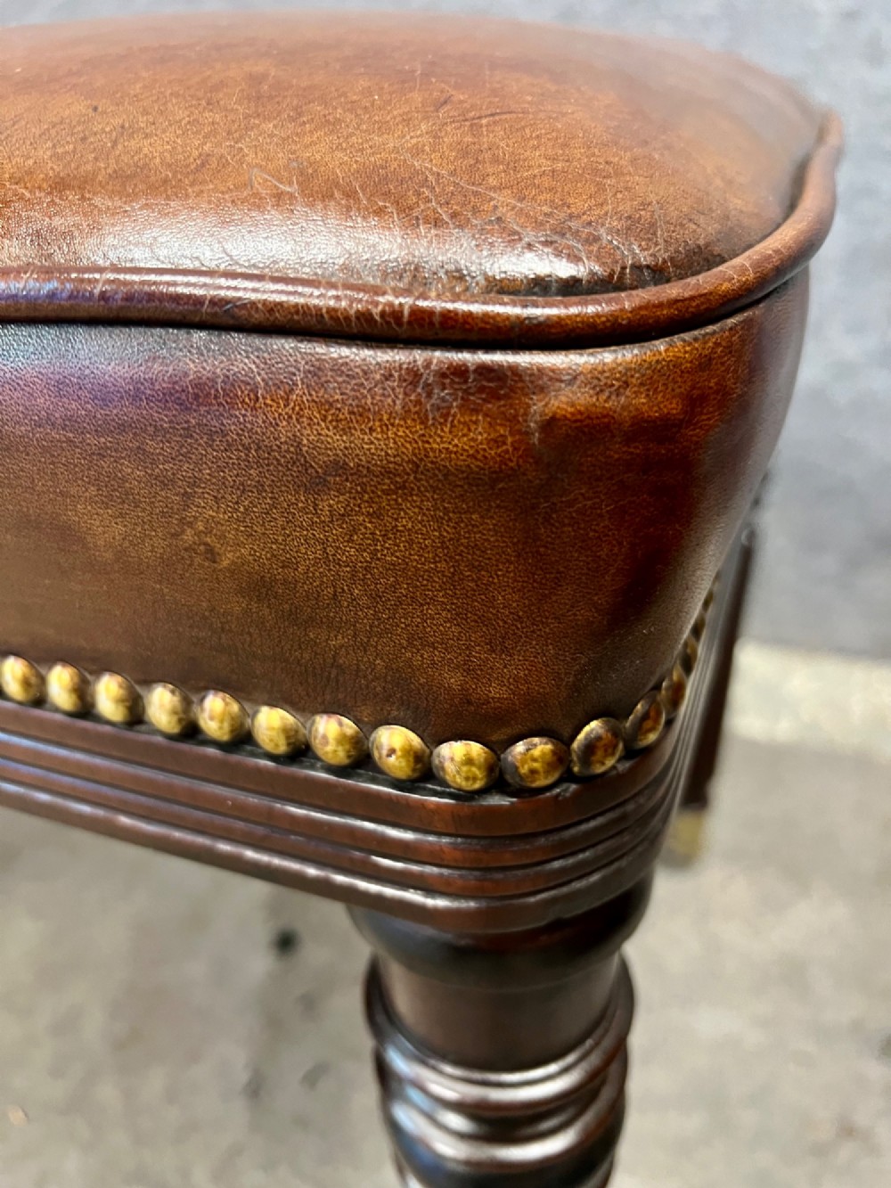 An early nineteenth-century upholstered brown leather bench featuring brass studded trim and six dark wood fluted turned legs on brass castors. View X
