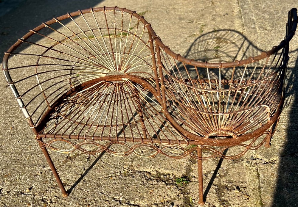 A weathered brown wrought iron S-shaped conversation seat featuring fan-patterned wire backrests and a looped apron base. View VII