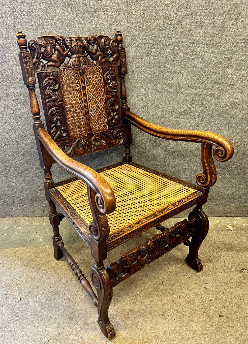 A dark carved oak Carolean style armchair featuring high cane back panels, a cane seat, scrolled armrests, and cherub crestings.