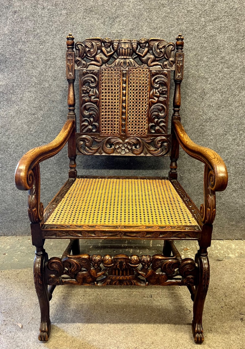 A dark carved oak Carolean style armchair featuring high cane back panels, a cane seat, scrolled armrests, and cherub crestings. View II