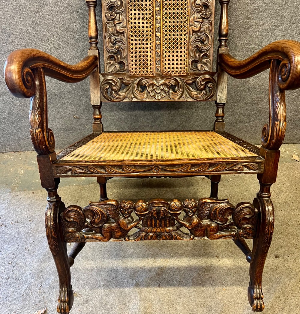 A dark carved oak Carolean style armchair featuring high cane back panels, a cane seat, scrolled armrests, and cherub crestings. View V