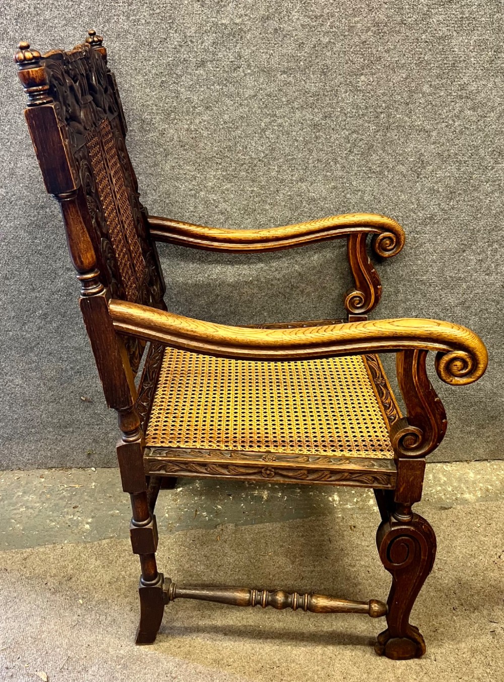 A dark carved oak Carolean style armchair featuring high cane back panels, a cane seat, scrolled armrests, and cherub crestings. View VII
