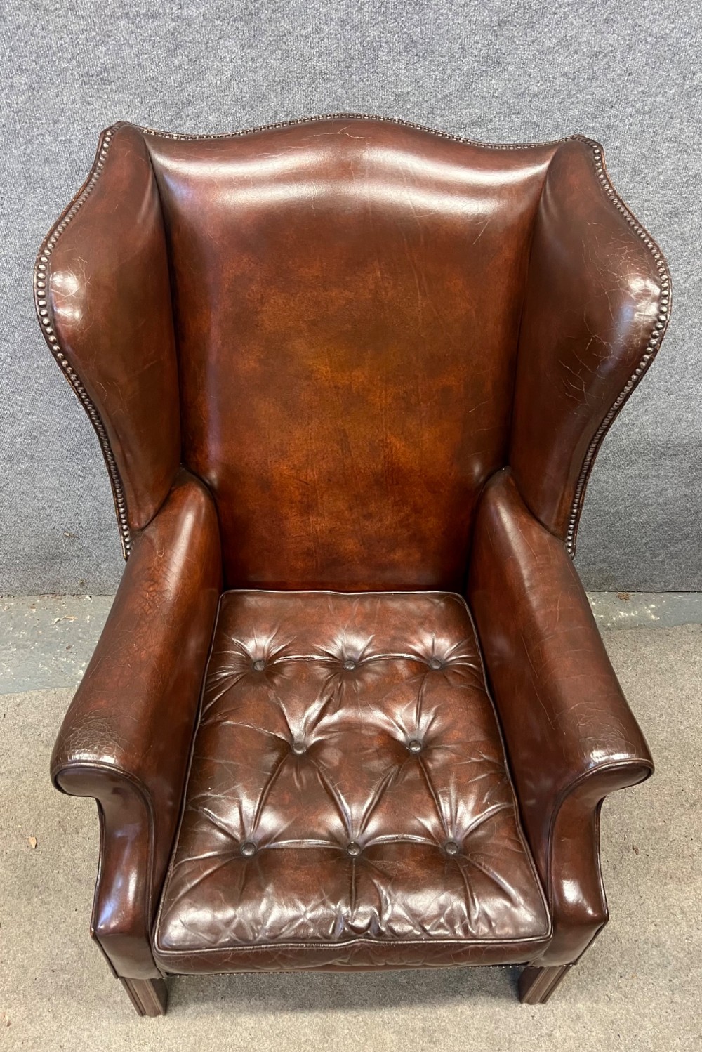 A Georgian style warm brown leather wingback armchair featuring a button-tufted seat cushion, brass stud trim, and dark timber legs. View II