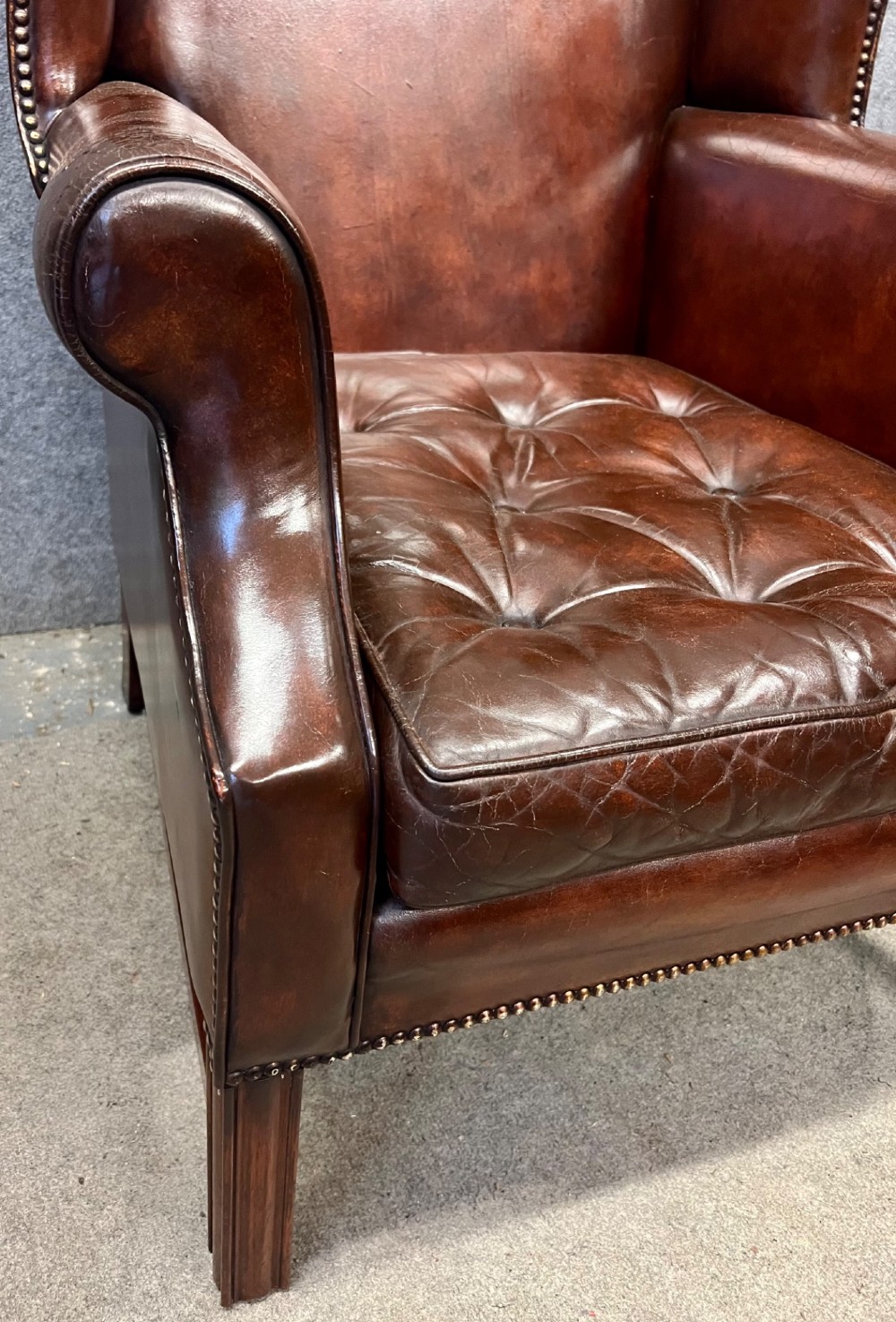 A Georgian style warm brown leather wingback armchair featuring a button-tufted seat cushion, brass stud trim, and dark timber legs. View V