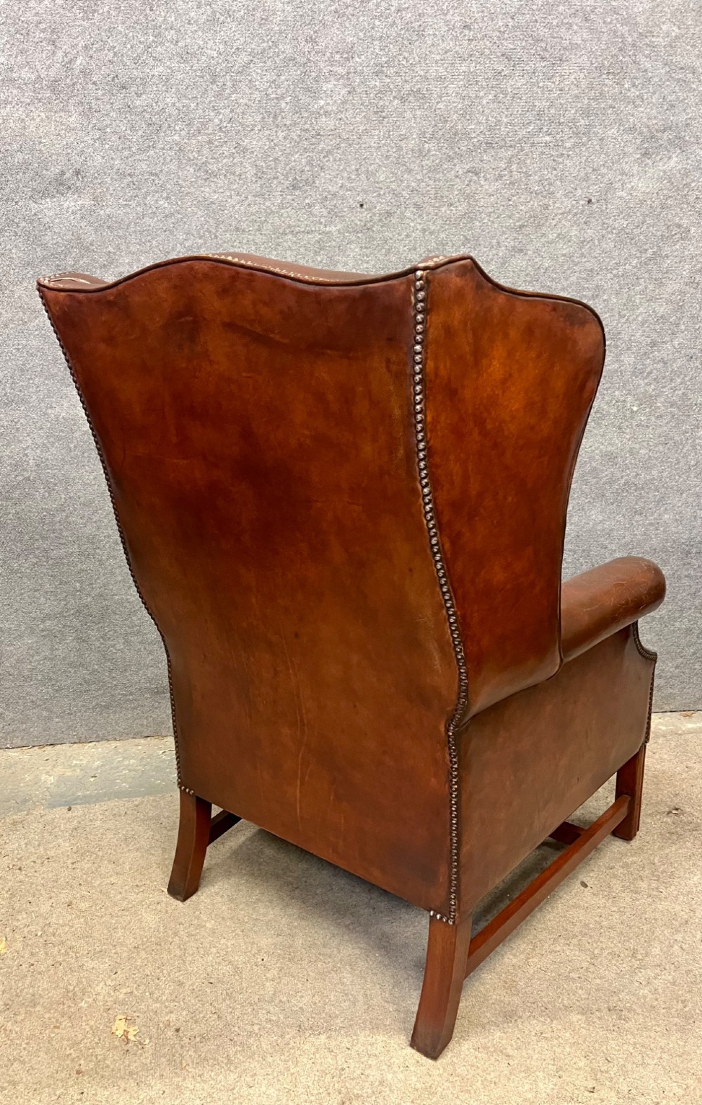 A Georgian style warm brown leather wingback armchair featuring a button-tufted seat cushion, brass stud trim, and dark timber legs. View VI
