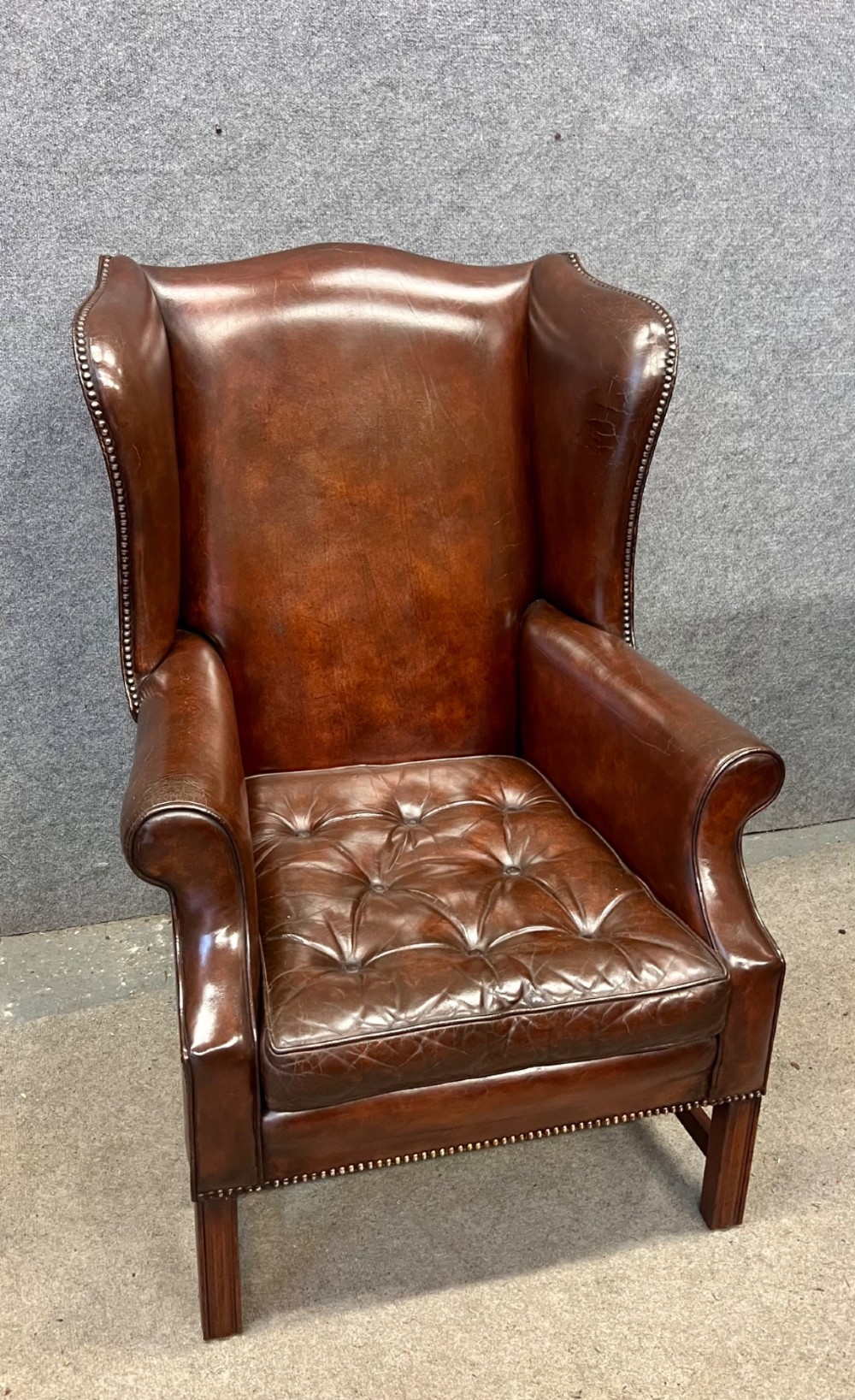 A Georgian style warm brown leather wingback armchair featuring a button-tufted seat cushion, brass stud trim, and dark timber legs. View IX