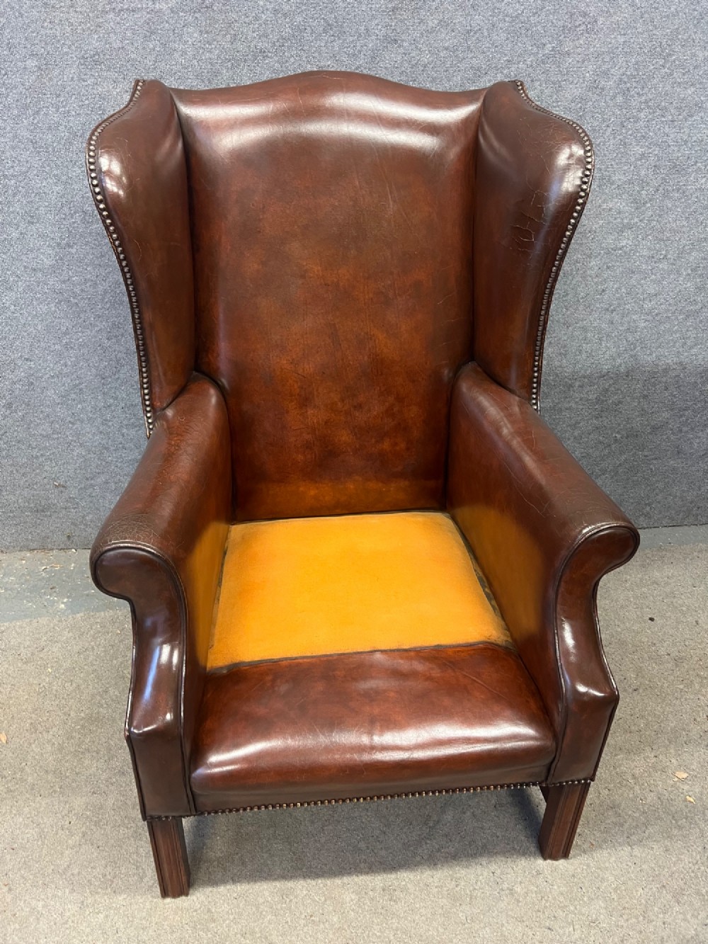 A Georgian style warm brown leather wingback armchair featuring a button-tufted seat cushion, brass stud trim, and dark timber legs. View X