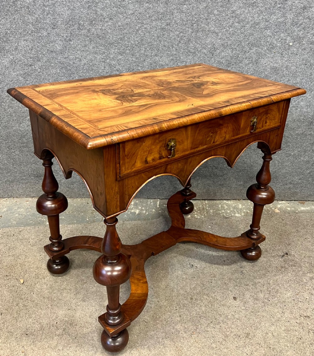 A William and Mary style walnut lowboy table featuring a quartered figured veneer top, a single drawer with brass drop handles, a shaped apron, turned legs, and a serpentine stretcher.