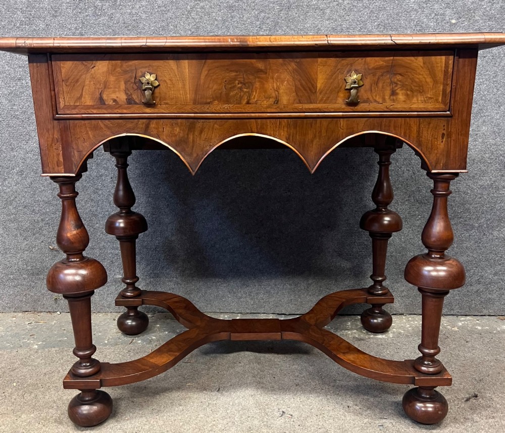 A William and Mary style walnut lowboy table featuring a quartered figured veneer top, a single drawer with brass drop handles, a shaped apron, turned legs, and a serpentine stretcher. View II